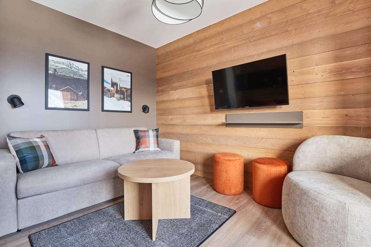 Ein modernes Wohnzimmer mit einer Couch, einem runden Couchtisch und orangefarbenen Hocker. Ein Flachbildfernseher ist an der Holzwand montiert.