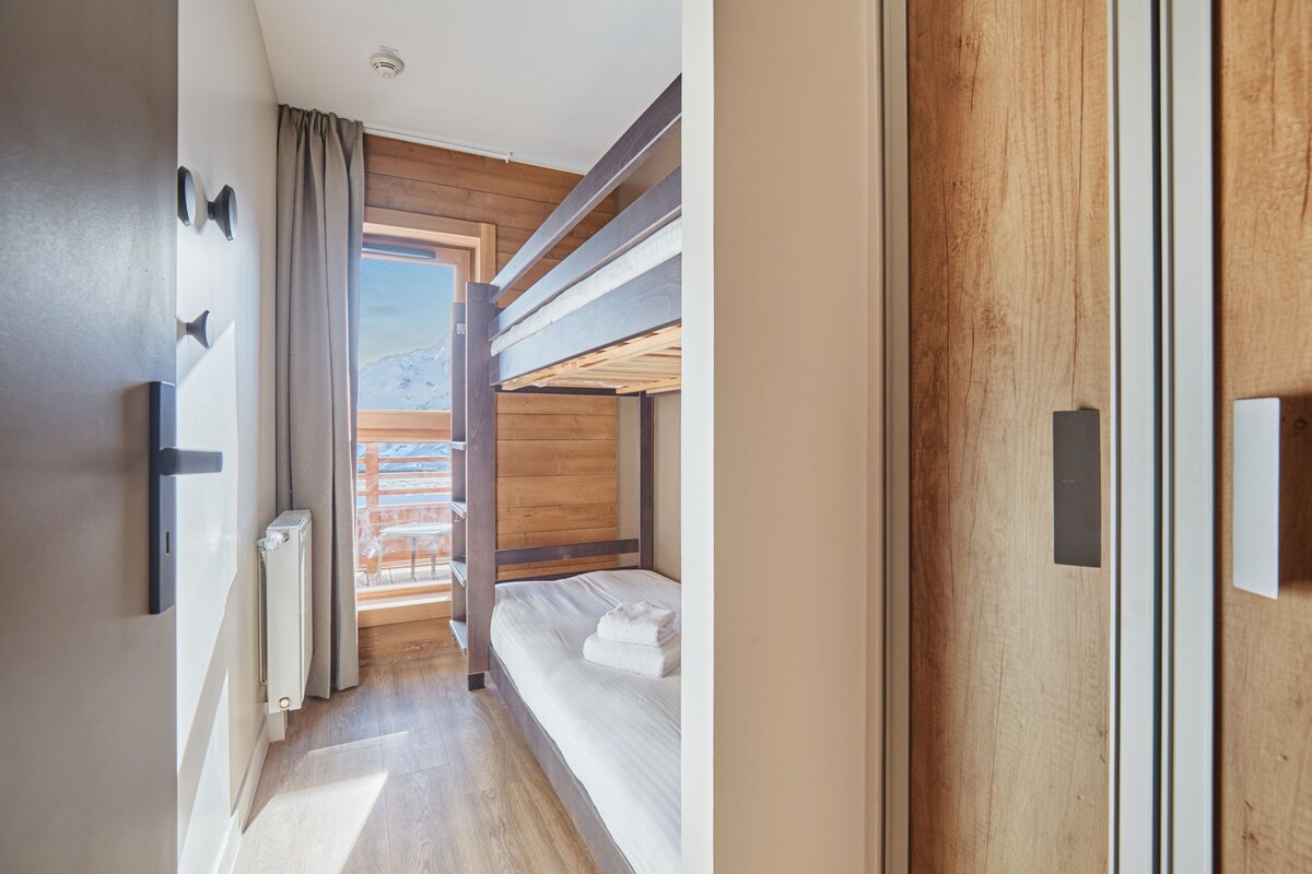A hotel room with a wooden floor, a bed with a white mattress, a window with a view of a wooden deck, and a door with a handle.