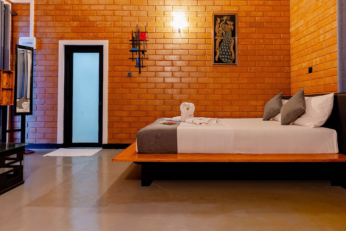 A hotel room with a brick wall, a bed with a white bedspread, two pillows, a wooden bench, a small table, a wall-mounted light, and a framed artwork.
