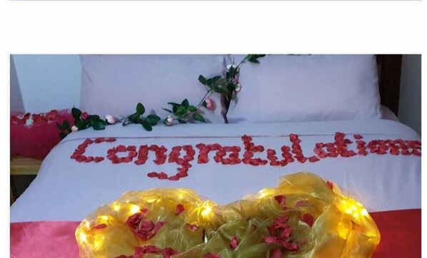 A bed with a white sheet, a garland of red roses, and a heart-shaped decoration made of rose petals and yellow lights.