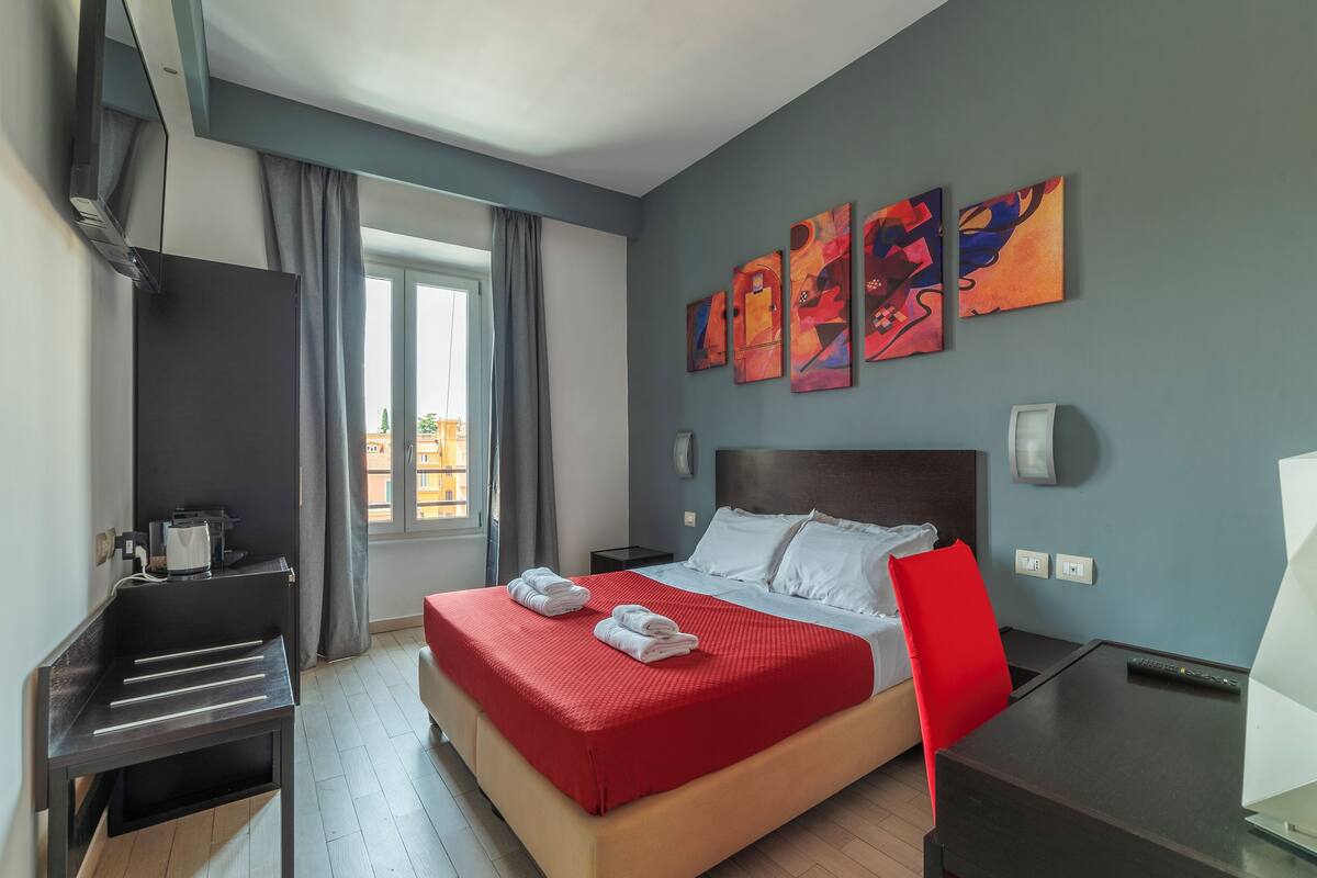 A hotel room with a bed, a desk, a chair, a window with curtains, and abstract wall art.