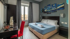 A hotel room with a bed, a desk with a computer, a chair, and artwork on the wall.