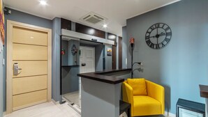 A modern living room with a yellow armchair, a clock on the wall, and a door leading to a kitchen area. - S. Marco Guesthouse (Rome)