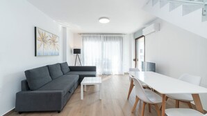 Un salon moderne avec un canapé gris, une table à manger blanche avec des chaises, un lampadaire et une grande fenêtre ornée de rideaux légers.