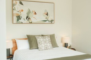 Ein ordentlich bezogenes Bett mit weißer Decke und zwei Kissen, eines gemustert und eines schlichtgrün, in einem Zimmer mit schlichter Wand und einem floralen Bild im Rahmen über dem Bett.