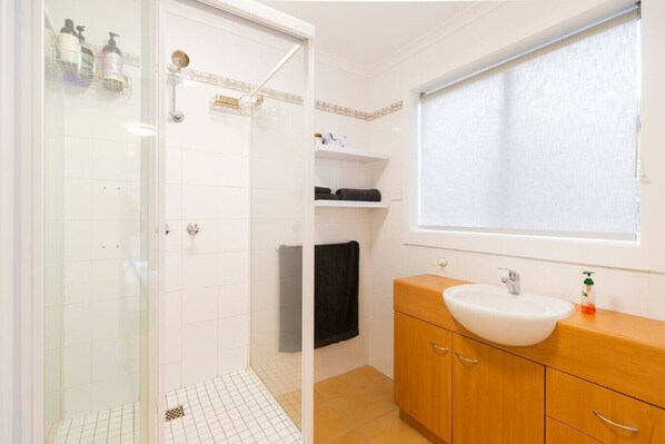 A bathroom with a glass shower enclosure, a sink with a wooden vanity, and a window with blinds.