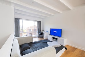 A modern bedroom with a bed, a desk, a TV, and a large window with curtains. - Renovated two-level Sandy Bay apartment (Sandy Bay)