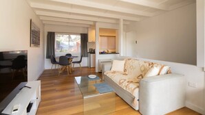 A modern living room with a sofa, a dining table with chairs, a kitchen area, and a view through a window. - Renovated two-level Sandy Bay apartment (Sandy Bay)