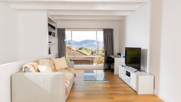 A modern living room with a sofa, TV, and a view of a cityscape through large windows. - Renovated two-level Sandy Bay apartment (Sandy Bay)