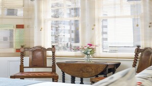 Ein Eckzimmer mit einem Holzbstuhl, einem kleinen Tisch mit Blumen in einer Vase und Blick durch große Fenster mit Jalousien.