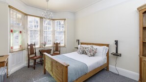 Ein Schlafzimmer mit einem hölzernen Bett, einem Schreibtisch mit Stuhl, einer Chandelier und großen Fenstern mit Jalousien.