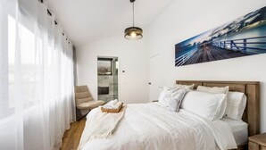 Une chambre à coucher avec un lit, une chaise, une fenêtre avec des rideaux légers, un tableau au mur et un luminaire au plafond.