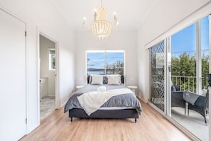 A bedroom with a large bed, a chandelier, a view of the outdoors, and a balcony with outdoor furniture.