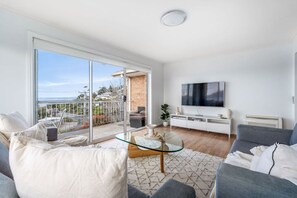 Ein modernes Wohnzimmer mit einem Glastisch, grauen Sofas, einem Flachbildfernseher und einem Balkon mit Blick.