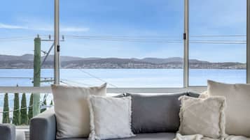 Quarto com sofá cinza e almofadas brancas, janelas amplas com vista para um corpo de água e terra ao longe.