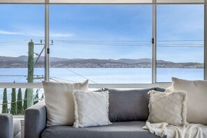 Ein Zimmer mit einer grauen Couch und weißen Kissen, große Fenster mit Blick auf ein Gewässer und entferntes Land.