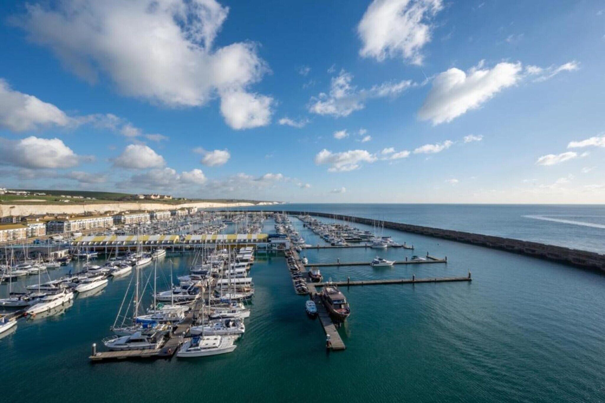 Un puerto deportivo con numerosas embarcaciones amarradas, un muelle que se adentra en el agua y edificios al fondo.