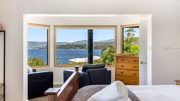 Ein Schlafzimmer mit einem großen Fenster, das Blick auf einen See und Hügel bietet. Es gibt ein Bett mit Holz Kopfteil, eine Kommode und zwei Stühle.