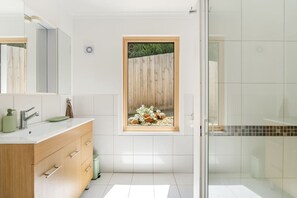 Een moderne badkamer met een grote spiegel, een houten wastafelmeubel, een raam met uitzicht op een houten schutting en planten, en een douchecabine met glazen wanden.