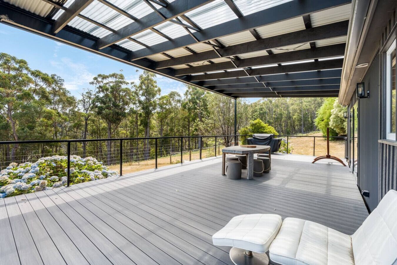 Une terrasse extérieure couverte, dotée d’une table et de chaises, entourée d’une rambarde en métal et offrant une vue sur les arbres.