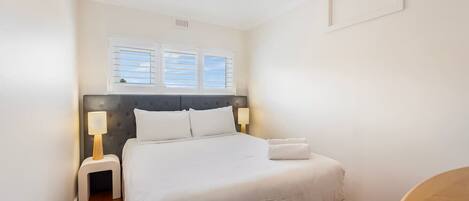 A neatly arranged bedroom with a large bed, two bedside lamps, and a window with blinds.