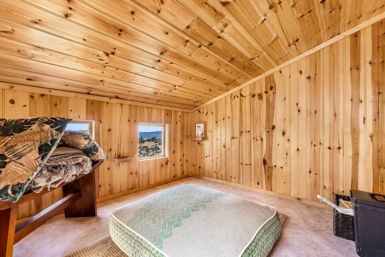 A wooden cabin room with a bed, a window with a view, and a small shelf with a framed picture.