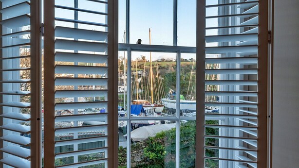 A view through a window with white blinds, revealing a marina with boats and a stone wall.