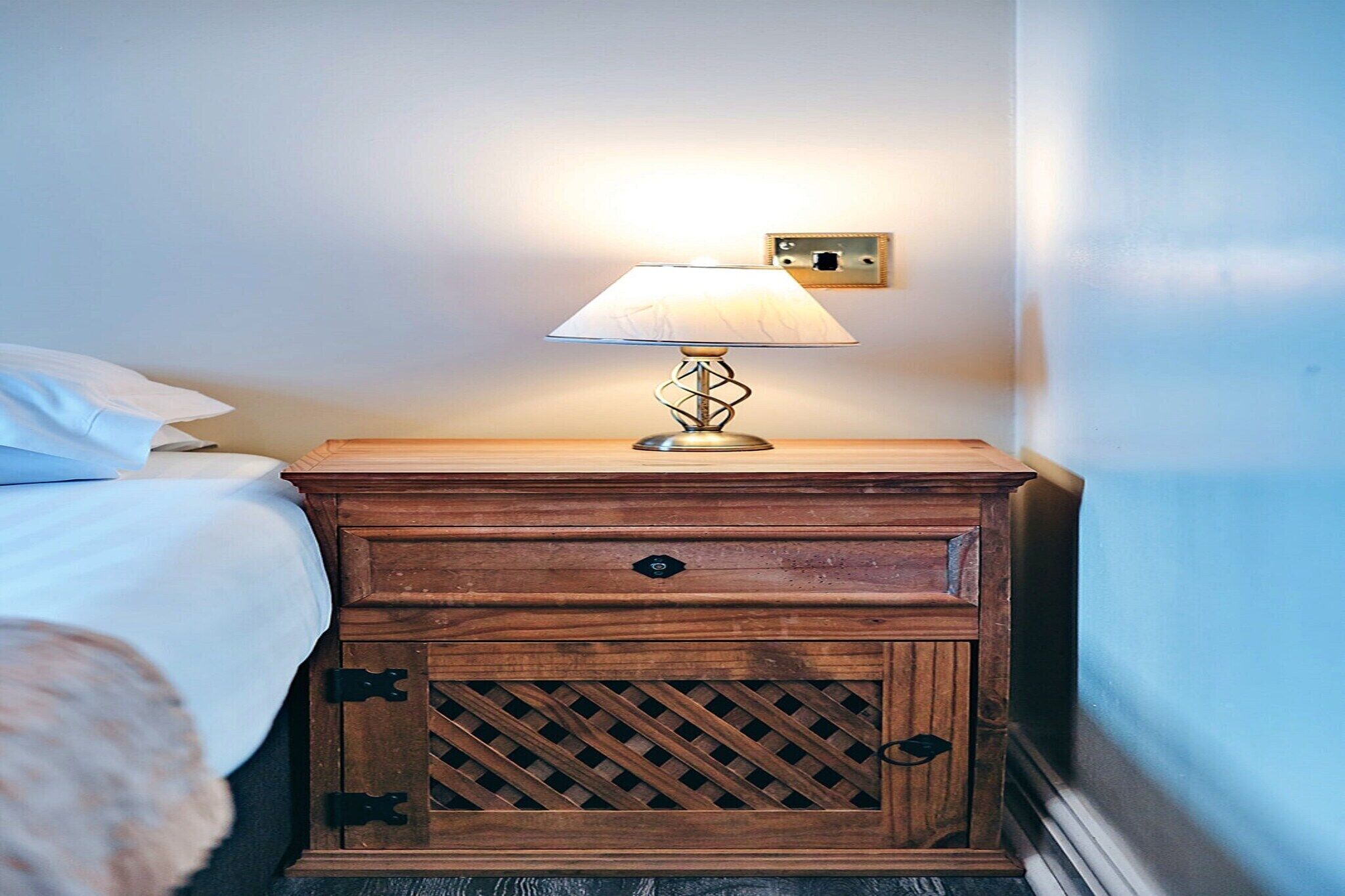 A wooden bedside table with a lamp, a bed with white bedding, and a wall socket.