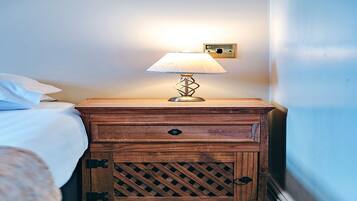 A wooden bedside table with a lamp, a bed with white bedding, and a wall socket.