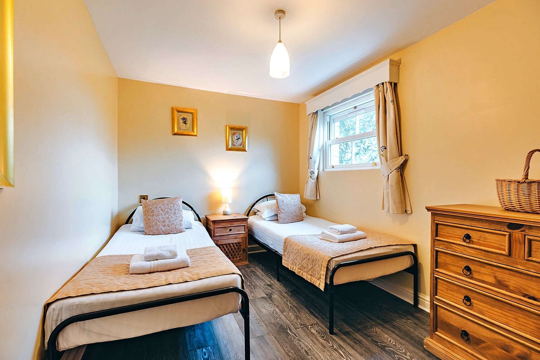 A room with two single beds, a wooden dresser, a window with curtains, and a hanging light fixture.