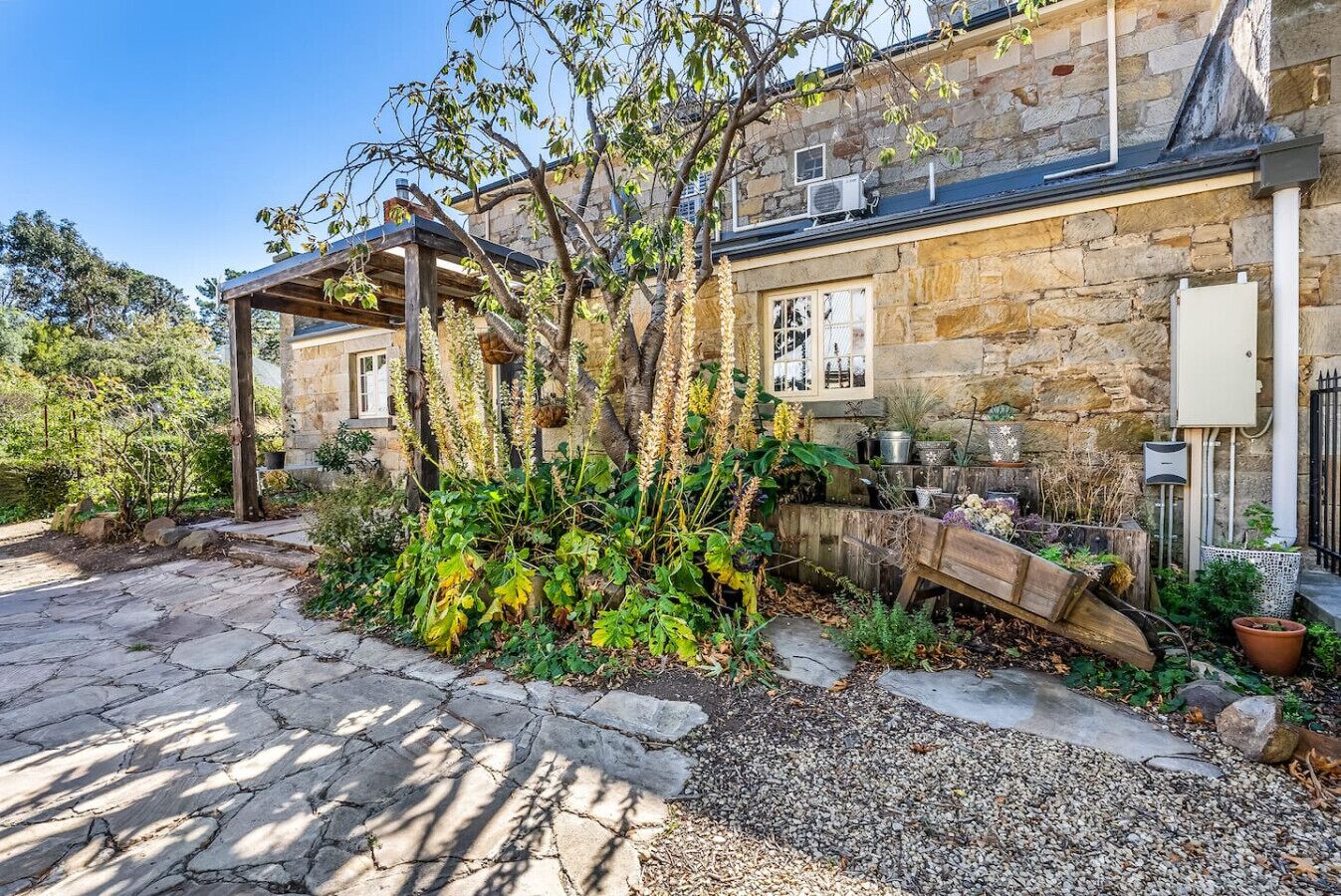 Un edificio de piedra con una pérgola de madera, un camino de grava y diferentes macetas con plantas.