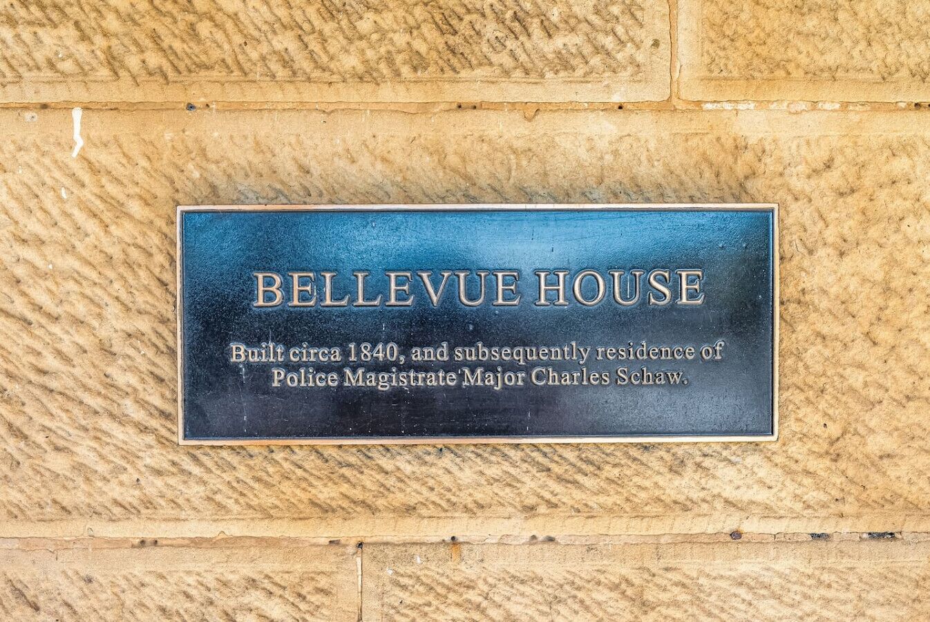 A plaque on a textured wall with the text 'BELLEVUE HOUSE Built circa 1840, and subsequently residence of Police Magistrate Major Charles Schaw.'