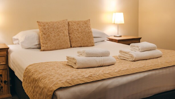 A neatly made bed with a beige bedspread, two matching pillows, and folded white towels on the bed. - Cottage in Netley Hall with Lush Gardens (Dorrington)