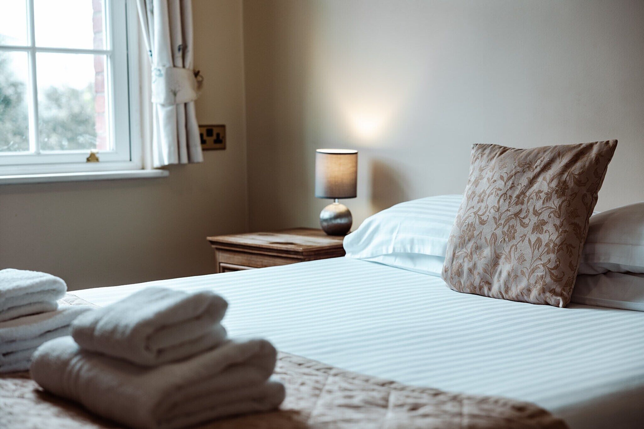 Een net opgemaakt bed met een kussen, een bedlamp en een raam met gordijnen.