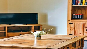 A wooden table with a vase of flowers in the center, a television on a stand, and a bookshelf in the background. - Cottage in Netley Hall with Scenic Trails (Dorrington)