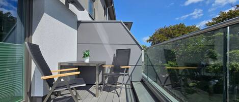Un balcón al aire libre con una mesa, dos sillas, una planta en maceta y barandilla de vidrio.