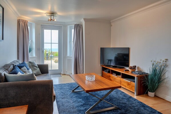 A modern living room with a sofa, a coffee table, a TV, and a wooden entertainment unit. - Apartment in Carbis Bay with Stunning Sea Views (St Ives)