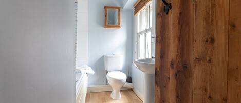 A narrow bathroom with a white toilet, a wooden door, and a window with blinds.