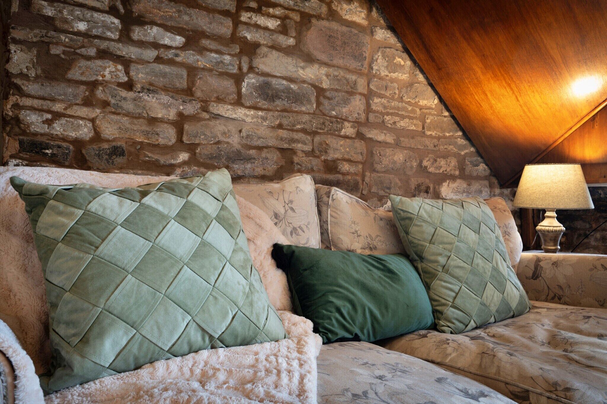 A cozy room with a stone wall and a wooden ceiling. There is a sofa with green and white cushions.