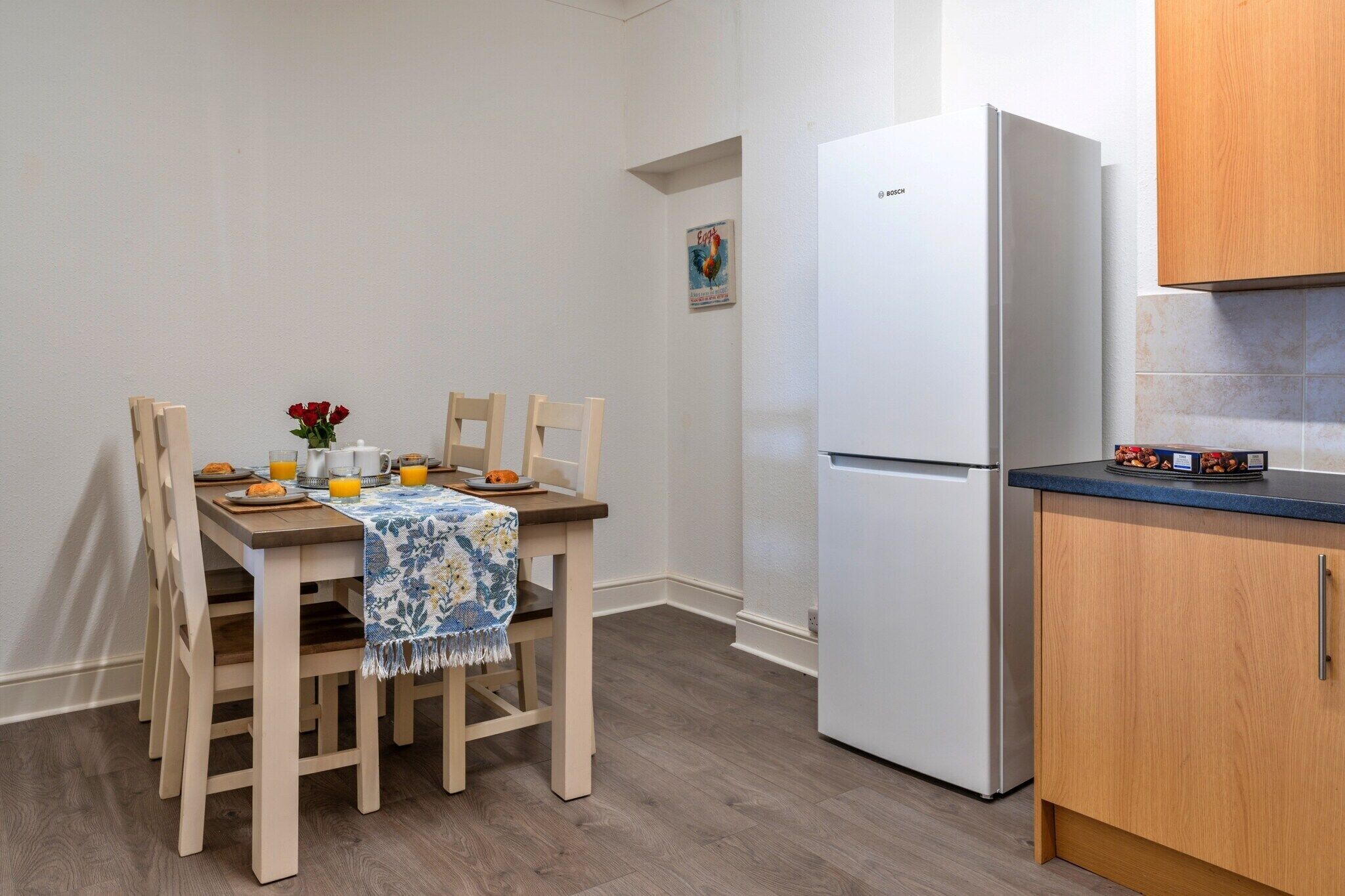 A small kitchen with a white refrigerator, wooden cabinets, a dining table with chairs, and a blue floral tablecloth.