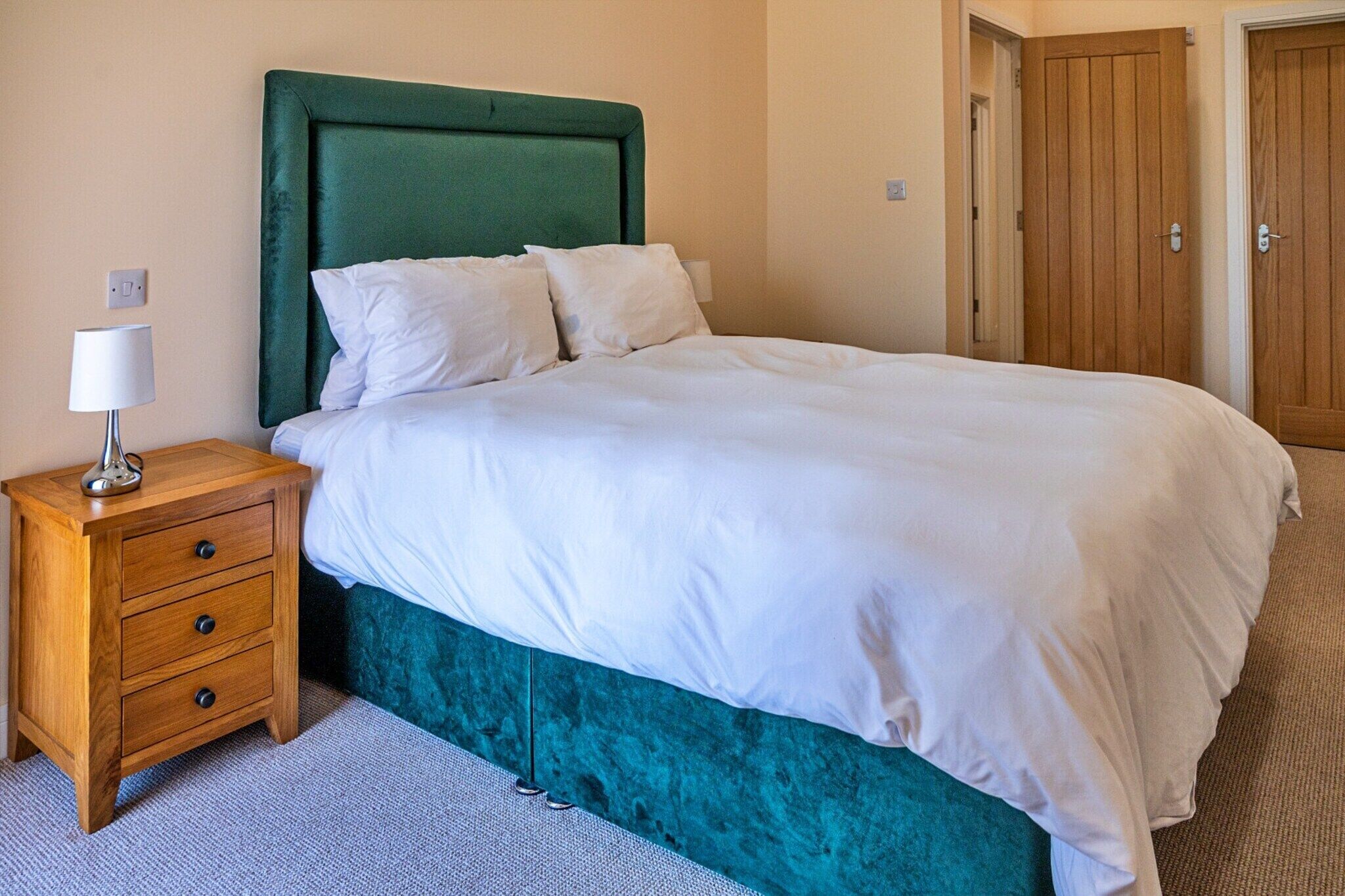 A neatly made bed with a green headboard, a wooden nightstand with drawers, and a lamp.