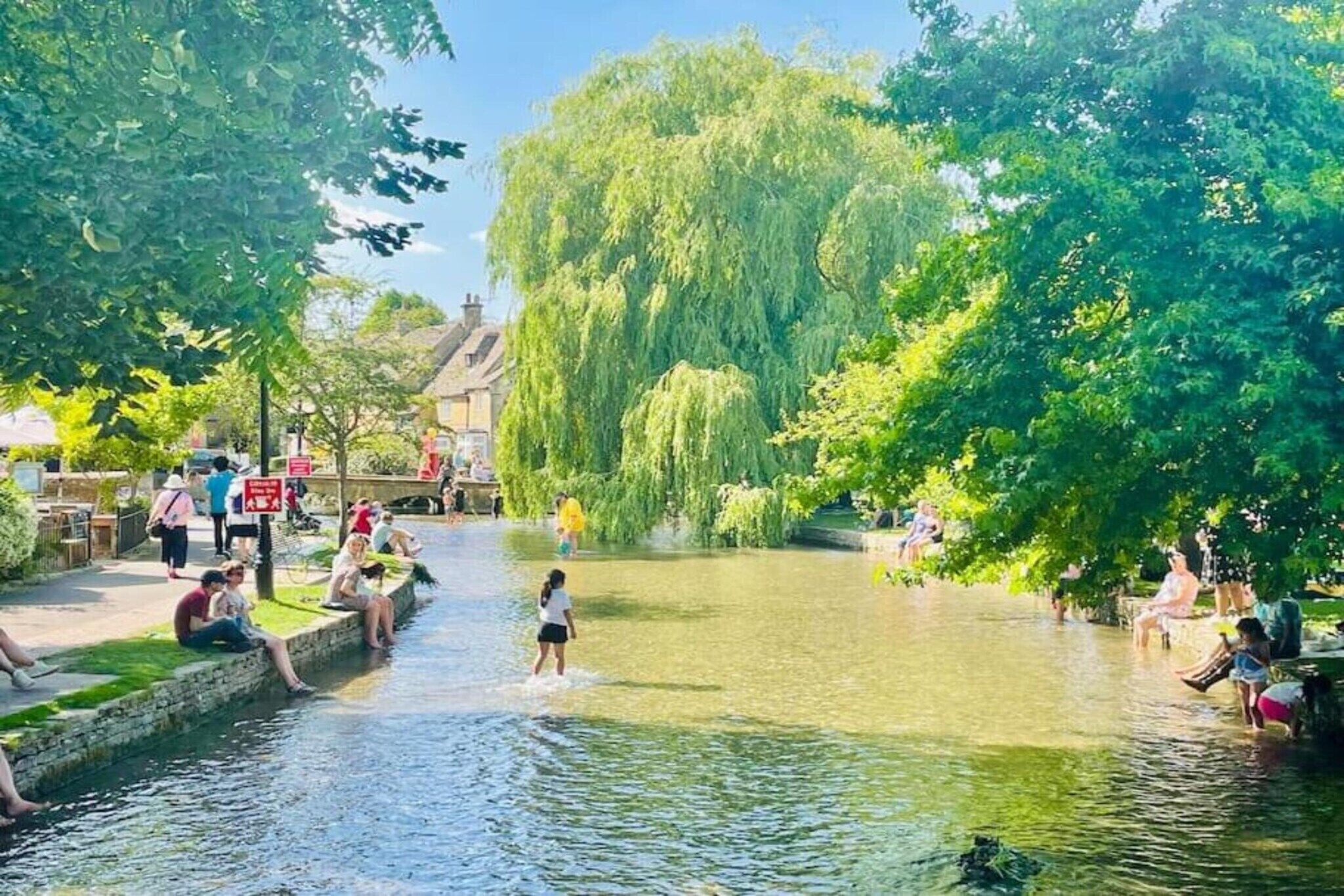 Ein Uferweg mit Menschen, die am Wasser entlanggehen und sitzen, umgeben von üppigem Grün und Bäumen.