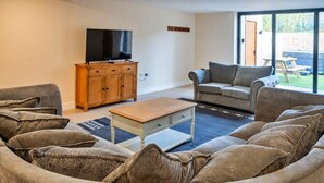 A living room with a sofa set, a coffee table, a TV on a wooden cabinet, and a sliding glass door leading to an outdoor area. - Barn in Cotswolds with Private Hot Tub (Little Rissington)