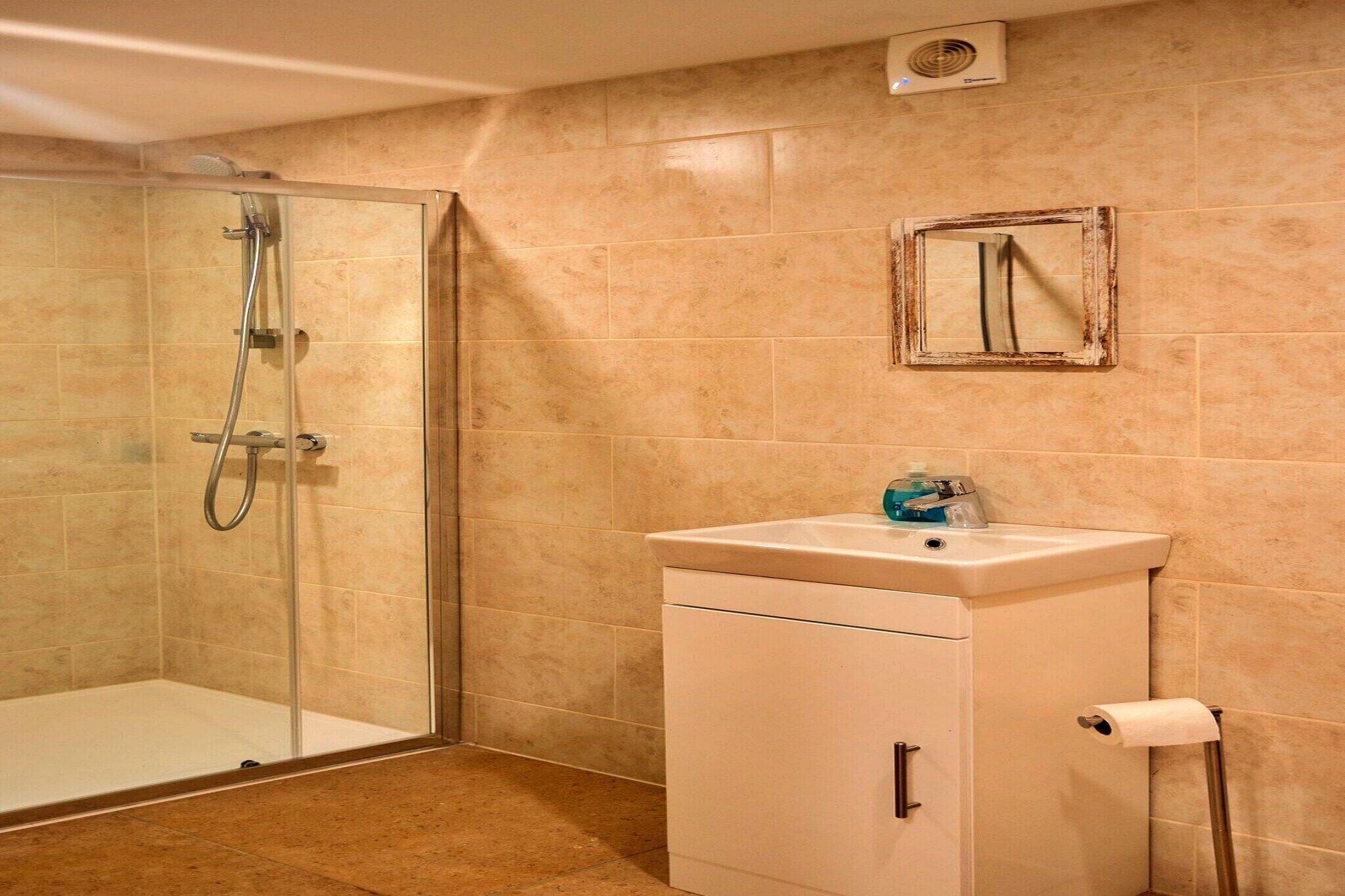 Een badkamer met een douchecabine van glas, een witte wastafel met spiegel erboven en beige tegels op de muren.