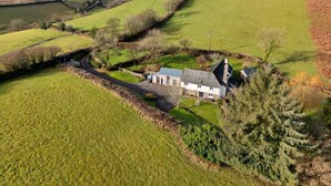 Une maison de campagne avec des fondations en pierre, entourée de champs verdoyants et bordée d’un chemin sinueux.