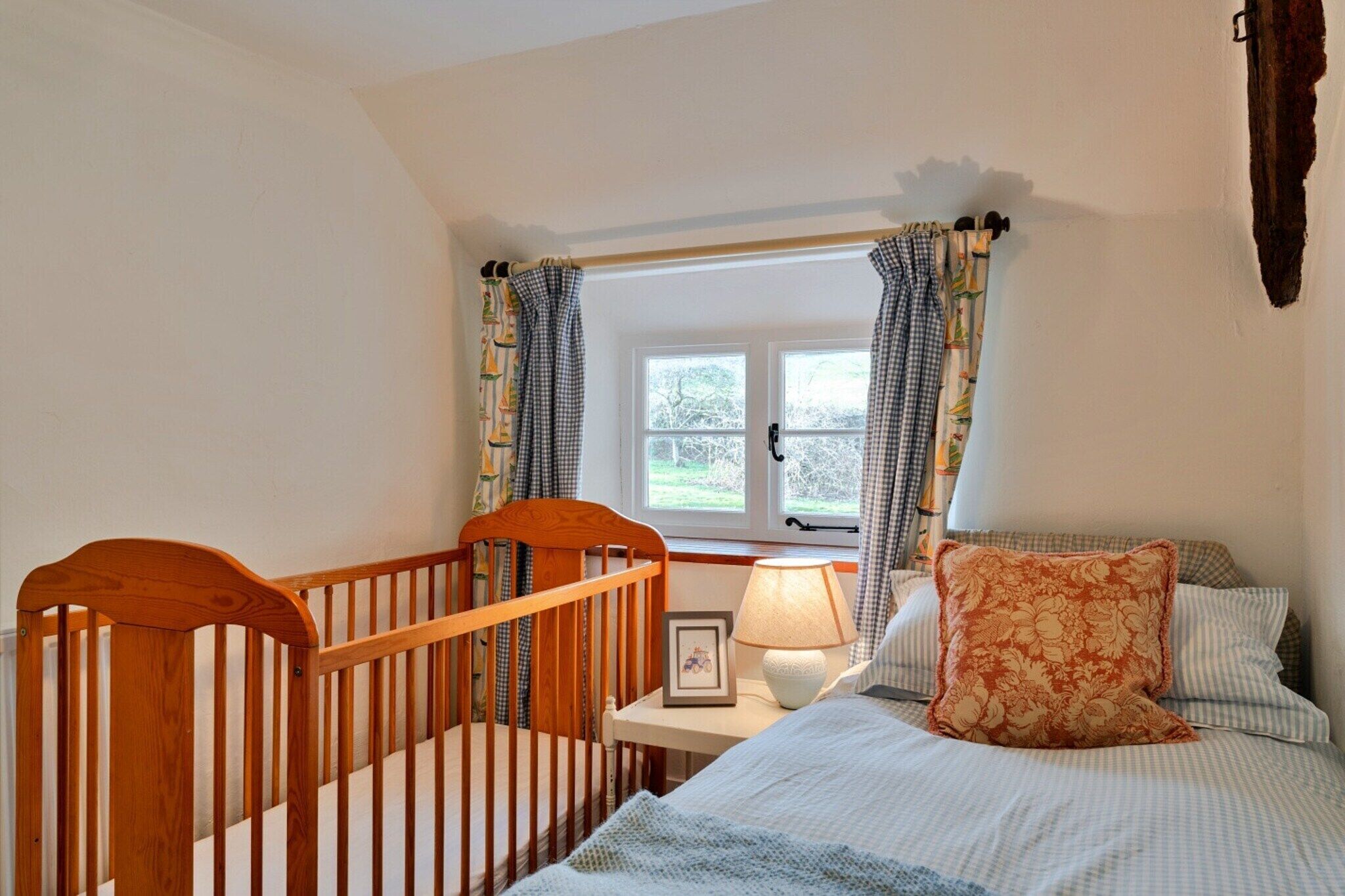 A bedroom with a crib, a bedside table with a lamp, a framed picture, and a bed with a floral pillow.