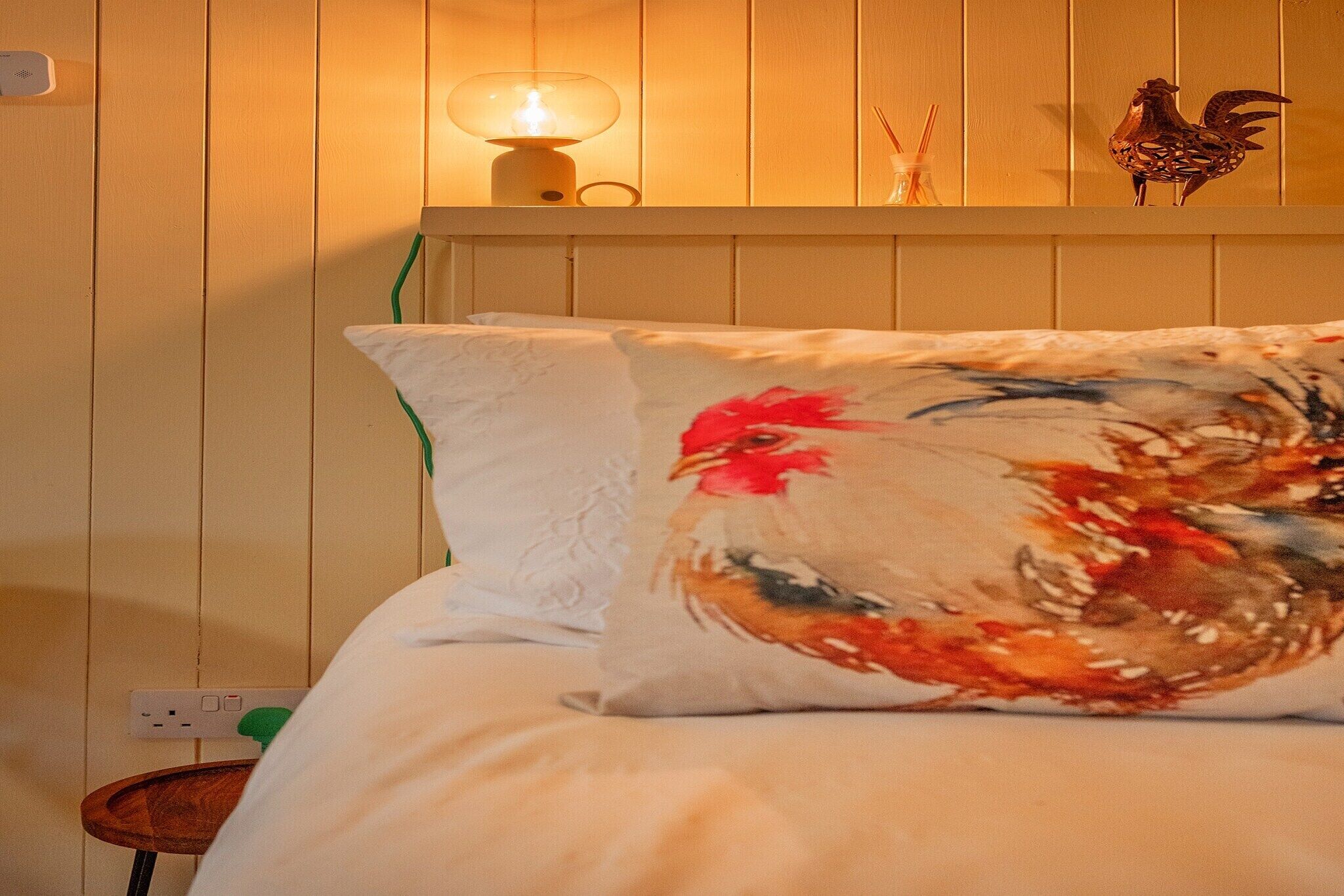 Una cama con un almohadón blanco y un cojín de colores con diseño de gallo.