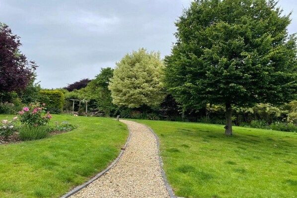 A garden pathway with a gravel edge, surrounded by lush green grass and various trees.