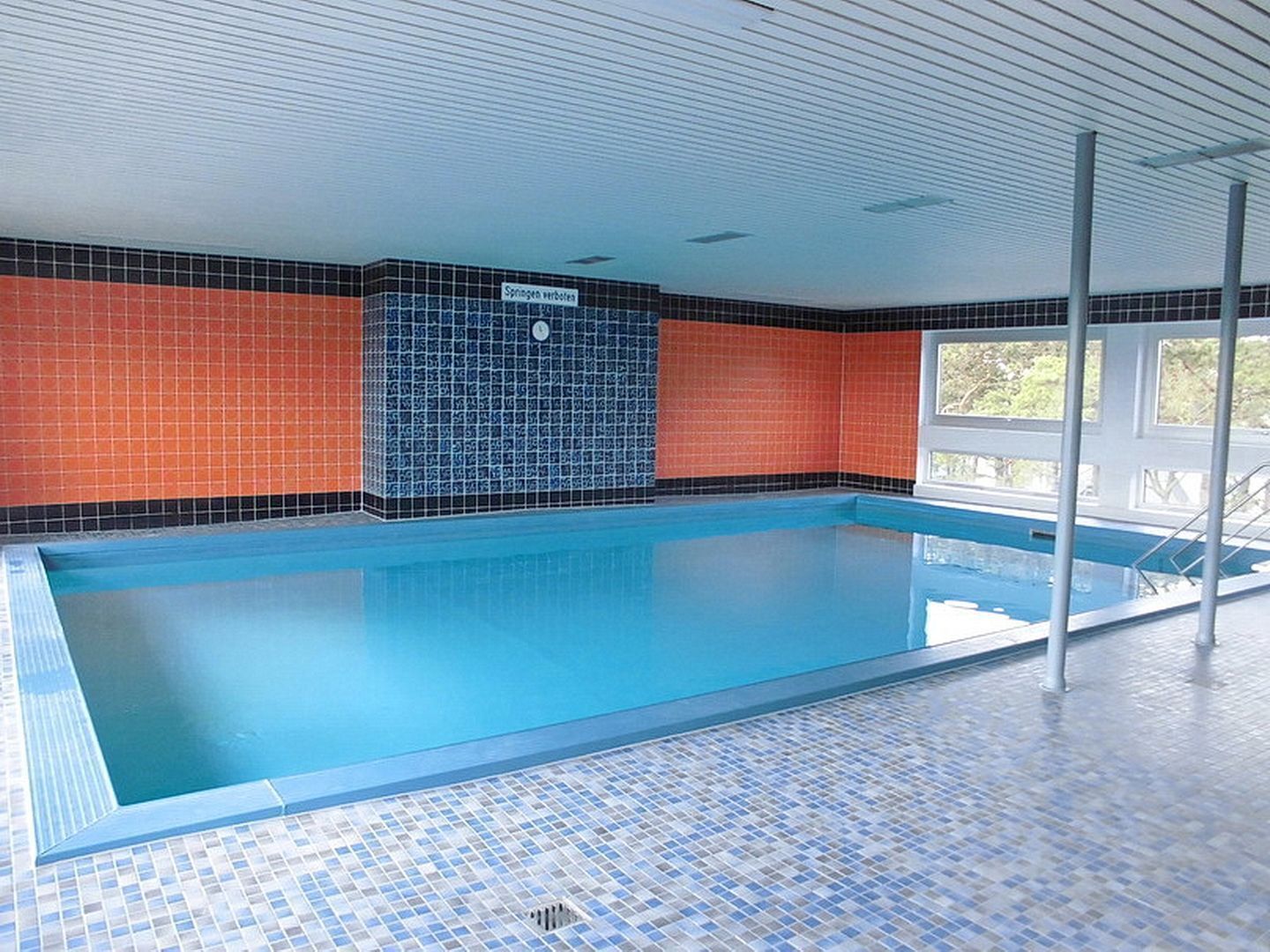 An indoor swimming pool with blue tiles, orange and black tiled walls, and a window on the right side.