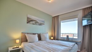 A hotel room with a large bed, a bedside table with a lamp, a flat-screen TV mounted on the wall, and a window with a view.
